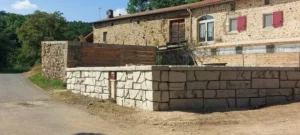 Mur de soutenement en pierre reconstituee bordant une cour, devant une ferme en pierre avec volets rouges et bardage bois