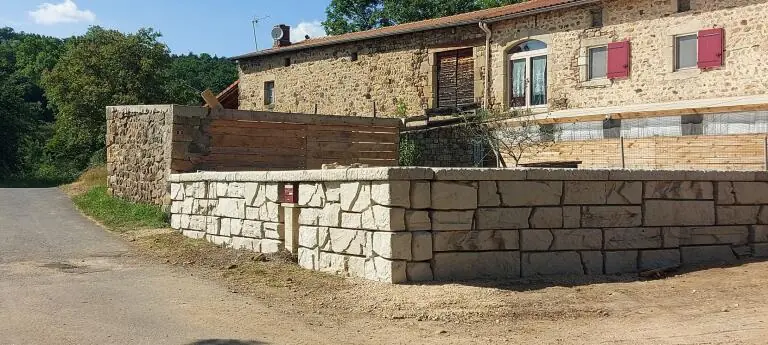 Mur de soutenement en pierre reconstituee bordant une cour, devant une ferme en pierre avec volets rouges et bardage bois