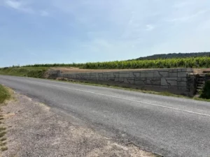 Long mur de soutenement en pierre le long d'une route, retenant une parcelle de vigne sous un ciel legerement nuageux