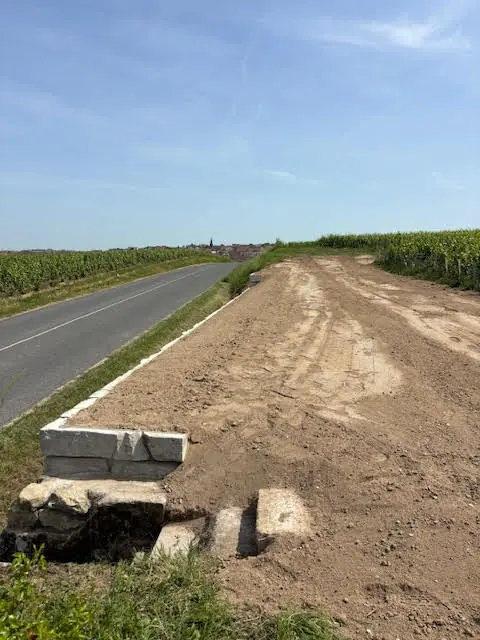Plateforme de terre nivelee en bord de route departementale, bordee de vignes, avec un village et son clocher a l'horizon